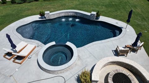 default A backyard pool area by Oklahoma Pool Builder featuring a uniquely shaped swimming pool and a connected circular hot tub. The poolside has several comfortable lounge chairs and umbrellas. Adjacent to the pool is a circular fire pit with seating. The surroundings are rich with green grass—a true custom pools oasis.