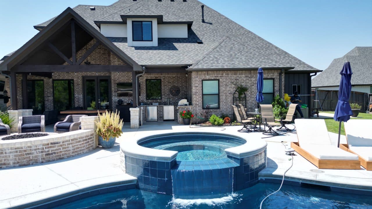 A backyard view of a house featuring a custom pool with an attached hot tub. The pool design includes lounge chairs, umbrellas, and a patio with seating and a grill. The house has a modern design with a gabled roof and brick exterior, set under a clear blue sky.