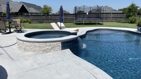 Anderson Pool A backyard with a Custom Pools design featuring a pool and hot tub, accompanied by lounge chairs. A trampoline is visible in the background, surrounded by a wooden fence, greenery, and neighboring houses. Two closed patio umbrellas stand by a table and chairs set. The sky is clear and blue.