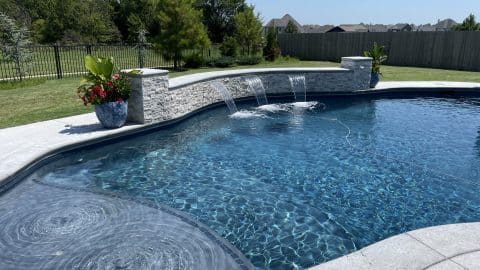 Anderson Pool A backyard swimming pool with clear blue water features three small waterfalls flowing from a white stone wall. Two potted plants with greenery and flowers sit on the wall's ends. Expertly crafted through thoughtful pool construction, the oasis is surrounded by a fence and lush, green lawn with trees in the background.