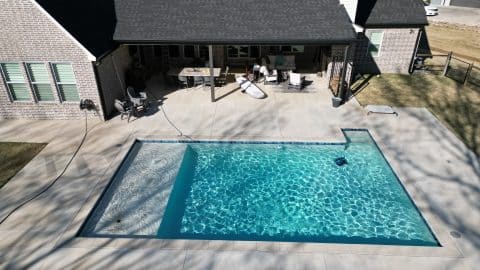 Aerial view of a residential backyard featuring a modern house with a dark roof and a large covered patio. The patio has outdoor furniture, a grill, and pool equipment. A rectangular swimming pool by Villa Del Mar Pools occupies the center of the concrete patio, casting clear reflections.