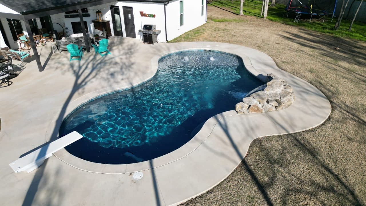 A backyard features a kidney-shaped swimming pool, expertly crafted by Villa Del Mar Pools, surrounded by a concrete deck. A diving board is at one end of the pool, and there's a small rock formation on one side of the water. The house has a patio with outdoor furniture, and a trampoline is visible in the background.