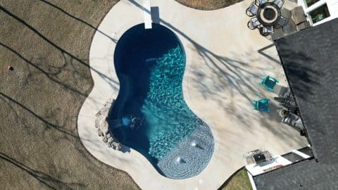 default Aerial view of a backyard featuring a custom kidney-shaped swimming pool by Villa Del Mar Pools, complete with a diving board and lounge chairs. Nearby, an outdoor patio with a round table and chairs invites relaxation. Adjacent to the pool is a house with part of the roof visible, shadows of trees falling gracefully on the area.