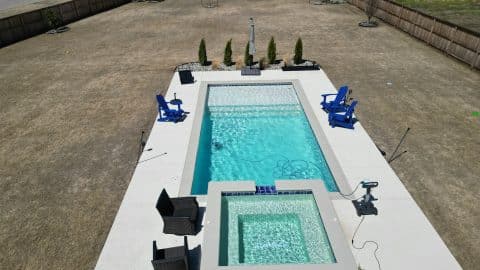 A backyard with a rectangular swimming pool and an attached smaller pool showcases impressive custom pool design. Four blue chairs line the pool's right side, and two black chairs sit near the smaller pool. The yard is enclosed by a wooden fence, with sparse vegetation and a few small trees planted along the perimeter.