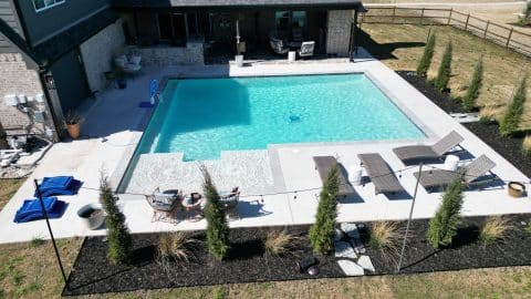 default A modern backyard features a sizable rectangular pool with clear blue water, showcasing the exceptional craftsmanship of an Oklahoma Pool Builder. Sun loungers and small tables are placed around the pool's edge. The area is surrounded by young trees and a black mulch border, with a spacious patio and house in the background.