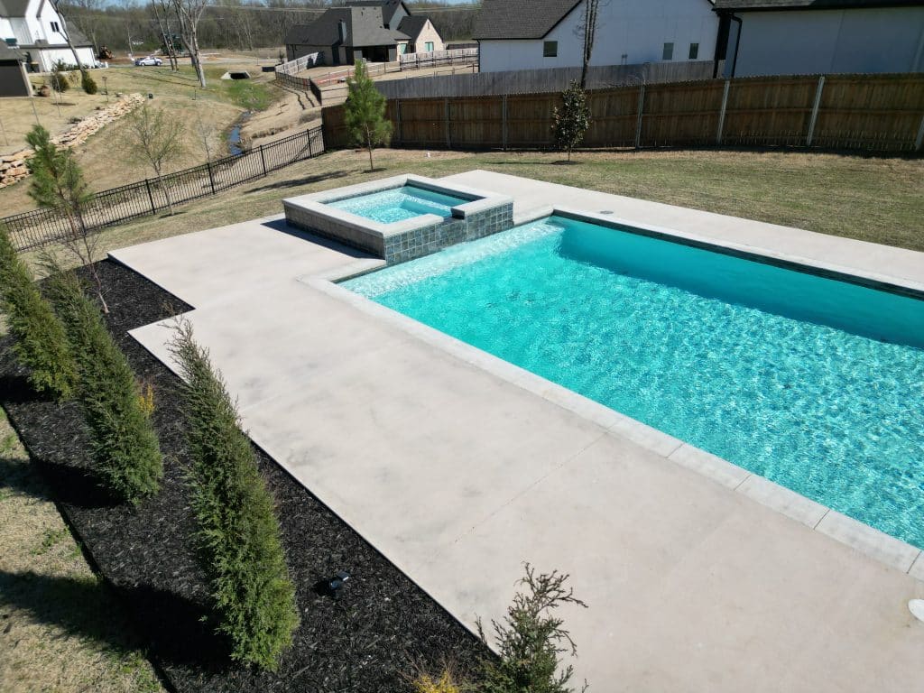 A backyard rectangular pool with a connected hot tub surrounded by a concrete patio. Designed by an expert Oklahoma Pool Builder, the area is bordered by young evergreens on black mulch. The yard includes a wooden privacy fence, and houses are visible in the background under a clear sky.