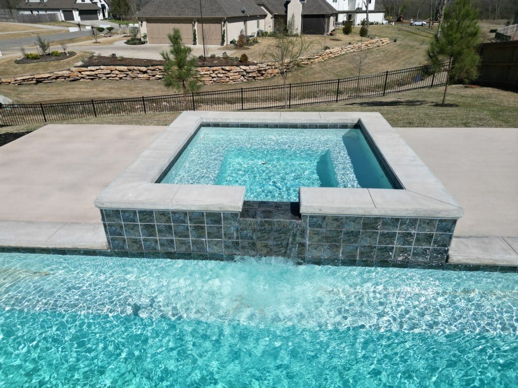 A luxurious outdoor pool with sparkling blue water features an elevated, rectangular hot tub with tile siding, spilling water into the pool below. Expertly crafted by an Oklahoma Pool Builder, it is set against a backdrop of houses and a stone retaining wall in a grassy landscape.