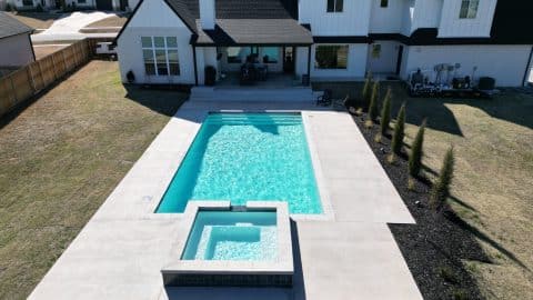 A backyard with a rectangular in-ground swimming pool and adjacent hot tub by Villa Del Mar Pools. The pool area, showcasing premium pool design, is surrounded by a concrete patio with garden beds and young trees. The main house, featuring a modern black and white design, stands prominently in the background.