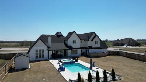 default A modern, two-story white house with black roof accents features a spacious backyard that includes a custom pool with a smaller spa section. The yard, surrounded by a wooden fence, boasts a grassy lawn with young trees planted near the pool area. It's perfect for anyone considering an Oklahoma Pool Builder.