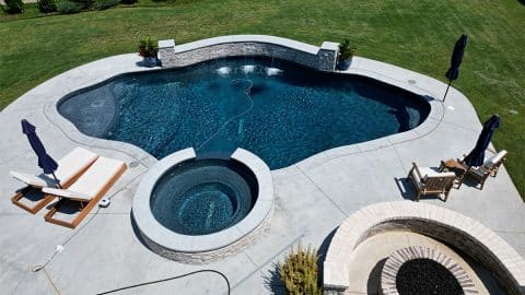 A backyard swimming pool with a circular hot tub attached showcases an elegant freeform design, brought to life by a reputable Oklahoma Pool Builder. Surrounded by a concrete deck, it features lounge chairs with umbrellas, a seating area with a fire pit, and a lush lawn enclosed by a fence.