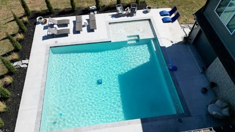 Aerial view of a backyard pool area with a large square pool and a smaller spa, designed by Oklahoma Pool Builder. Lounge chairs and umbrellas are arranged on the concrete deck. The area, crafted by Villa Del Mar Pools, is surrounded by a lawn and a wooden fence. A house is partially visible on the right side.