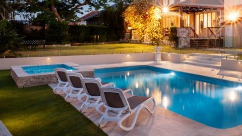 A luxurious backyard features a glowing custom pool with a built-in hot tub, surrounded by a lush lawn and various plants. Four lounge chairs are placed poolside. In the background, there is a well-lit two-story house with a spacious porch and large windows, showcasing impeccable pool design.