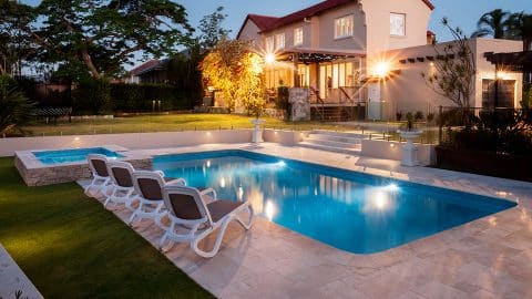 villadelmar process graphic A luxurious house with a well-lit exterior at dusk features a sparkling rectangular swimming pool, custom-designed by an Oklahoma Pool Builder. Four lounge chairs beside the pool and a manicured lawn create an inviting space while trees and shrubs surround the property, enhancing the serene ambiance.