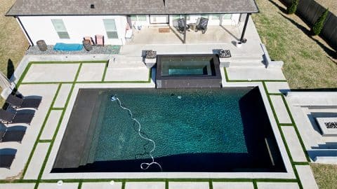 villadelmar tyler graphic Aerial view of a modern backyard featuring a rectangular swimming pool with steps and a submerged ledge, crafted by the expert team at Oklahoma Pool Builder. The pool area includes lounging chairs, an adjacent hot tub, and a well-maintained lawn framed by concrete pathways. A house is visible in the background.